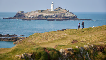 Grodrevy lighthouse This landscape photograph captures Godrevy Lighthouse, the main subject, situated on a rocky island off the coast of Cornwall, United Kingdom. The image was taken in the early afternoon during spring, reflecting the clear visibility and gentle light typical of that season. The lighthouse, a prominent coastal landmark, stands above rugged cliffs and is surrounded by the blue waters near Sennen Cove. Nature is evident in the grassy foreground and the coastal scenery, with two people and a dog visible walking along the cliffs. The scene highlights the natural beauty of Cornwall's coastline, with Godrevy Lighthouse serving as an iconic structure along this stretch of the United Kingdom's coast.